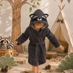 May include: A dark gray hooded bathrobe featuring a raccoon face design on the hood, complete with black and white ears, a black eye mask, and a small nose. The robe includes long sleeves, a matching tie belt at the waist, and two front pockets.