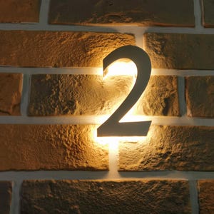 May include: A backlit metal number 2 mounted on a brick wall. The number is dark, and the light behind it creates a warm glow. The bricks are a mix of brown and tan.