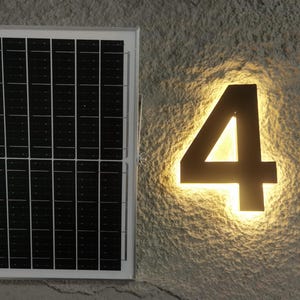 Puede incluir: Un panel solar con un patrón de cuadrícula está montado junto al número cuatro retroiluminado. El número es oscuro con un brillo cálido que emana de detrás, montado en una pared gris texturizada. El panel solar está en un marco blanco.
