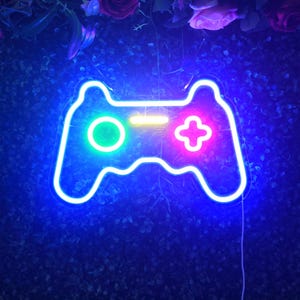 May include: A neon sign in the shape of a video game controller. The controller is outlined in bright blue, with a green circle on the left, a pink plus sign on the right, and a yellow bar in the center.