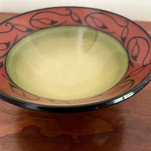 May include: A round ceramic bowl featuring a smooth, light green interior basin. The wide, flat rim is colored in a deep terracotta red and decorated with a continuous black line art pattern of stylized leaves and vines, finished with a black edge.