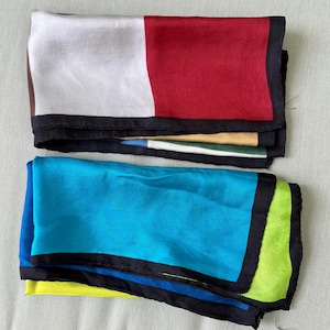 May include: Two folded square scarves with black borders. The top scarf features a white and red color-blocked design with hints of yellow and green underneath. The bottom scarf is bright blue with a lime green corner and a dark blue layer beneath.