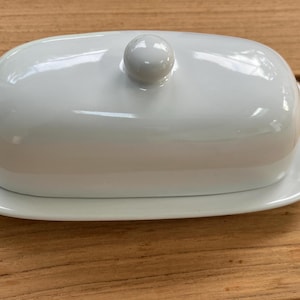 May include: A white ceramic butter dish with a smooth, glossy finish. The rectangular base features rounded corners and a matching domed lid with a small, round knob handle centered on top. The design is simple, clean, and functional.