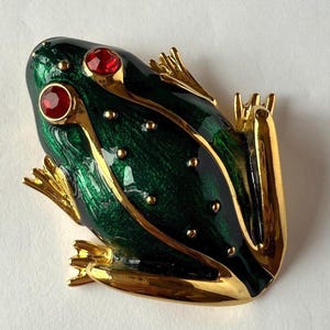 Peut inclure: Broche en forme de grenouille verte et dorée avec des yeux en pierres précieuses rouges. La broche présente un design détaillé avec des accents dorés et de petits points dorés. Le corps de la grenouille est en émail vert foncé.