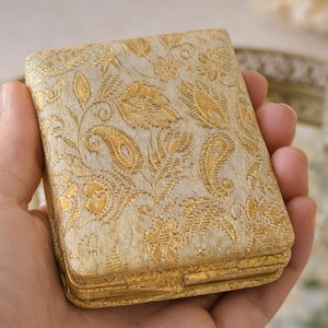Peut inclure: Un étui rectangulaire doré avec un motif cachemire orné. L'étui mesure un peu plus de 7,6 cm de long. L'étui est tenu dans une main, avec un plateau miroir et un flacon de parfum en arrière-plan.