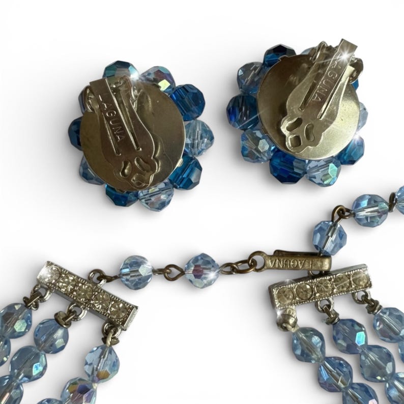 May include: A vintage blue beaded jewellery set, including a necklace and clip-on earrings. The earrings have a flower design with blue beads. The necklace features multiple strands of blue beads and a silver clasp. The earrings and necklace are marked "LAGUNA".