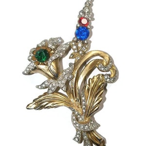 May include: A gold-toned brooch with a floral design. The brooch features a cluster of flowers with green, red, and blue rhinestones. The flowers are surrounded by smaller rhinestones.