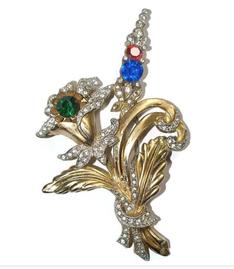 May include: A gold-toned brooch with a floral design. The brooch features a cluster of flowers with green, red, and blue rhinestones. The flowers are surrounded by smaller rhinestones.