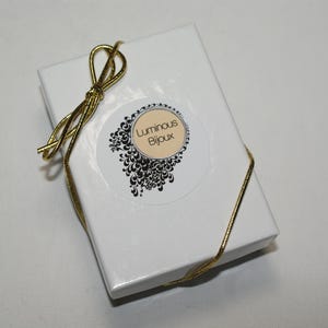 May include: A white gift box tied with a gold ribbon. The box has a circular sticker with the text "Luminous Bijoux" in a decorative font, surrounded by a black and white floral design.