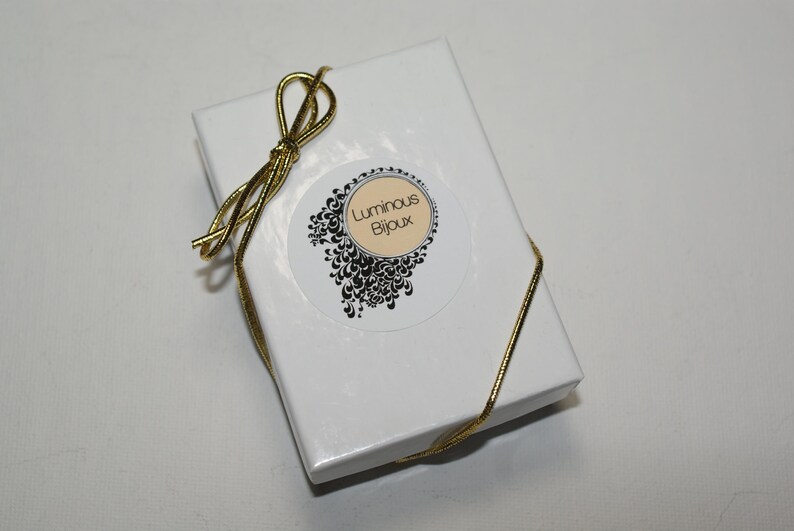 May include: A white gift box tied with a gold ribbon. The box has a circular sticker with the text "Luminous Bijoux" in a decorative font, surrounded by a black and white floral design.