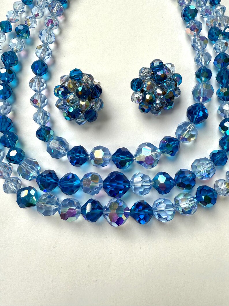 May include: A vintage jewellery set featuring a multi-strand beaded necklace and matching clip-on earrings. The beads are faceted and range in shades of blue, from light to deep sapphire, with iridescent accents. The earrings are clustered with similar beads.