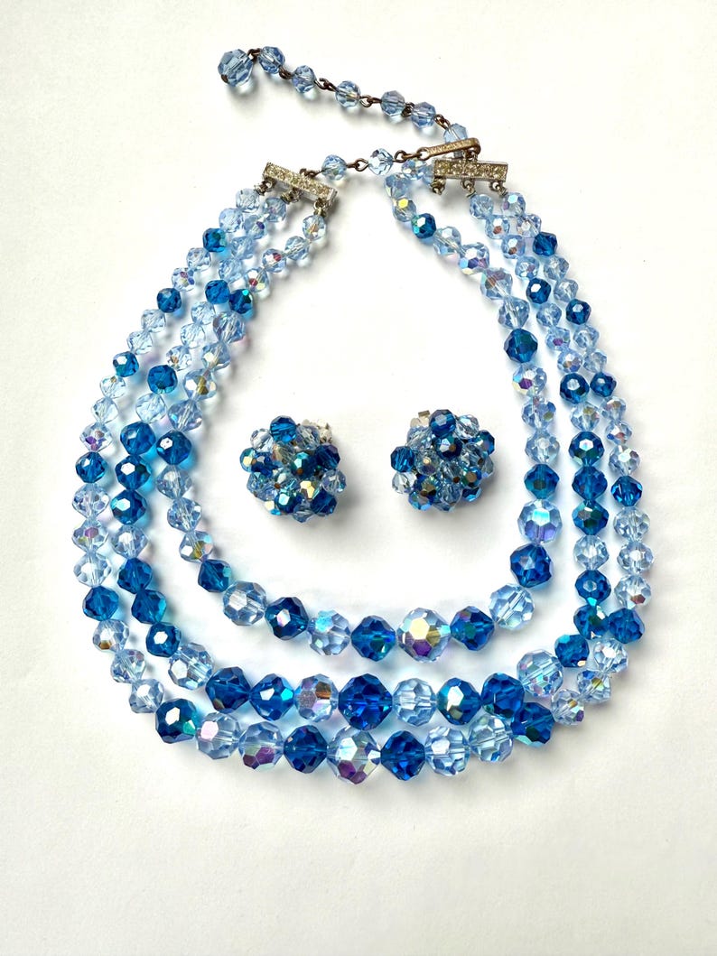 May include: A vintage jewellery set comprising a three-strand necklace and matching clip-on earrings. The necklace and earrings are made of faceted blue crystal beads in varying shades. The necklace has a silver-tone clasp.