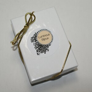 May include: A white gift box with a gold ribbon tied in a bow. The box has a round sticker with a black and white design and the text "Luminous Bijoux".