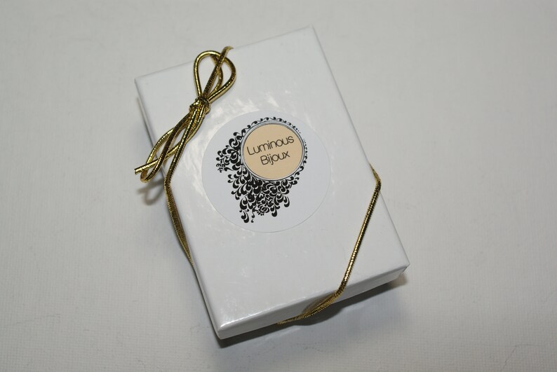May include: A white gift box with a gold ribbon tied in a bow. The box has a round sticker with a black and white design and the text "Luminous Bijoux".
