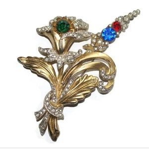 May include: A gold-tone brooch with a floral design featuring three flowers with rhinestones and colored stones. The flowers are green, blue, and red.