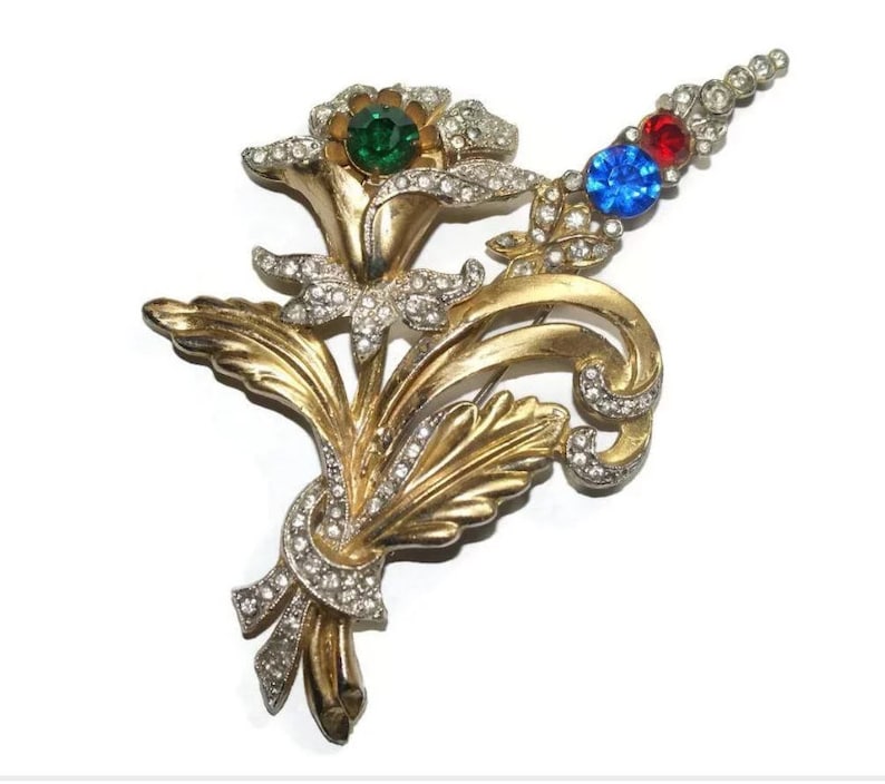 May include: A gold-tone brooch with a floral design featuring three flowers with rhinestones and colored stones. The flowers are green, blue, and red.