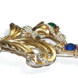 May include: A gold-toned brooch with a floral design, featuring a cluster of rhinestones and colorful stones in shades of green, blue, and red.
