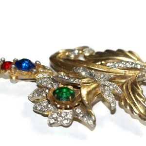 May include: A gold-tone brooch with a floral design, featuring red, blue, and green rhinestones. The brooch is made of metal and has a delicate, intricate design.