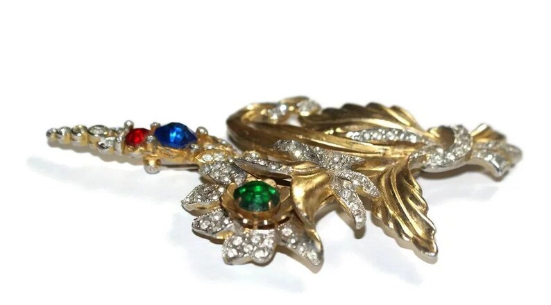 May include: A gold-tone brooch with a floral design, featuring red, blue, and green rhinestones. The brooch is made of metal and has a delicate, intricate design.
