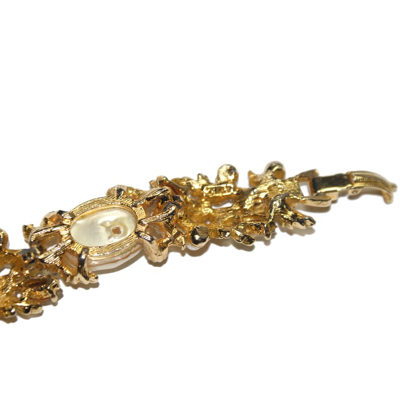 May include: A gold-tone bracelet with a large, oval-shaped faux pearl in the center. The bracelet is intricately designed with a floral pattern and has a clasp closure.