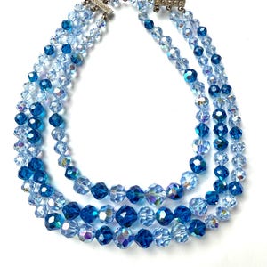 May include: A three-strand necklace with faceted blue crystal beads. The necklace features varying shades of blue, from light to deep, with a silver clasp. The beads are arranged in a graduated design, creating a layered effect.