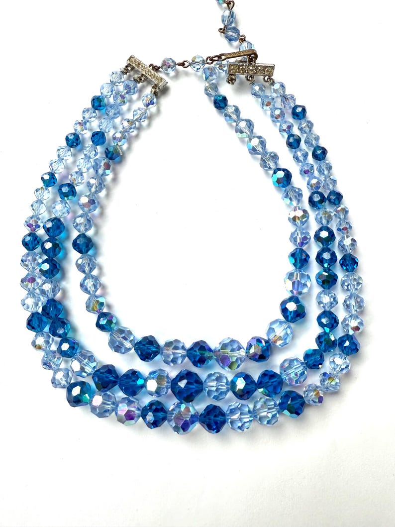 May include: A three-strand necklace with faceted blue crystal beads. The necklace features varying shades of blue, from light to deep, with a silver clasp. The beads are arranged in a graduated design, creating a layered effect.