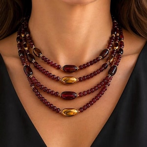 May include: A multi-strand necklace featuring four layers of deep red, faceted beads. Each strand is accented with a single, smooth, elongated central bead in alternating metallic gold and dark translucent red tones, creating a layered effect.