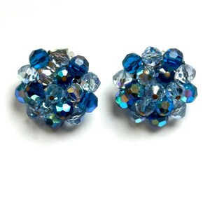 May include: A pair of vintage clip-on earrings. Each earring features a cluster of faceted beads in shades of blue, including deep navy, light blue, and iridescent clear beads. The beads are arranged in a spherical shape, creating a sparkling accessory.