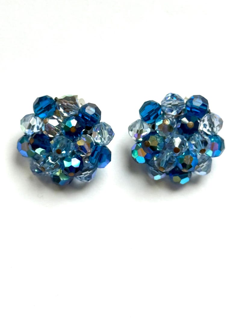 May include: A pair of vintage clip-on earrings. Each earring features a cluster of faceted beads in shades of blue, including deep navy, light blue, and iridescent clear beads. The beads are arranged in a spherical shape, creating a sparkling accessory.