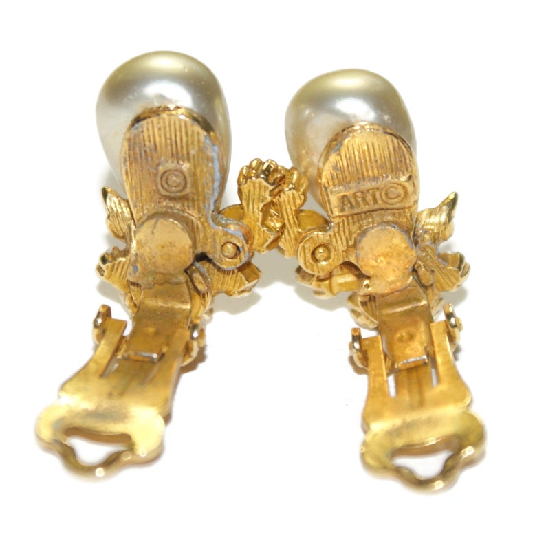 May include: A pair of gold tone clip-on earrings with faux pearl accents. The earrings feature a floral design and are marked "ARTIC" on the back.