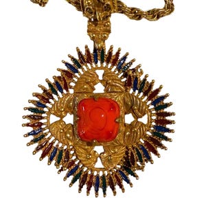 May include: Gold-toned pendant necklace with a large, square, orange center stone. The pendant features a sunburst design with red, blue, and green accents. The necklace has a thick chain.