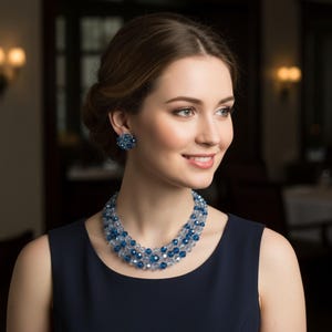 May include: A woman wearing a three-strand necklace and matching earrings made of blue and clear crystals. The jewellery is set against a dark blue dress. The woman is smiling, with a soft background of a room with warm lighting.