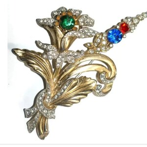 May include: A gold-toned brooch with a floral design. The brooch features a large green stone surrounded by smaller clear stones, and a smaller blue stone and red stone on a stem.