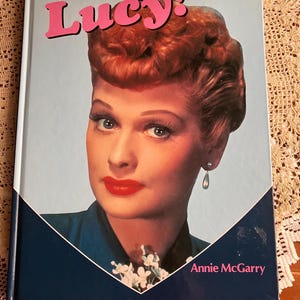 May include: A book titled "Lucy!" features a portrait of Lucille Ball with red hair, red lipstick, and a blue dress. The title is in pink letters above her image, set against a light blue background within a heart shape.