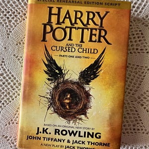 May include: A yellow and black book cover with the title "Harry Potter and the Cursed Child - Parts One and Two". The cover features a black bird with its wings spread, a nest, and a child inside the nest.