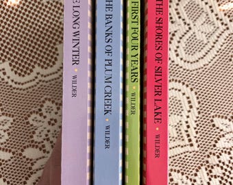 May include: Four hardcover books by Wilder are displayed vertically. The spines are in shades of purple, blue, green, and red. The titles include "The Long Winter", "On the Banks of Plum Creek", "The First Four Years", and "By the Shores of Silver Lake".