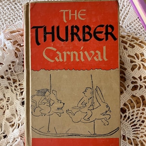 May include: Vintage hardcover book titled "The Thurber Carnival." The cover features a red and tan design with black lettering and illustrations of figures on a carousel. The book is set against a white lace background.