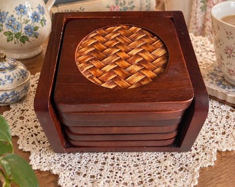 May include: A set of square dark brown wooden coasters stacked inside a matching wooden holder. Each coaster features a circular center inlay made of woven light brown rattan material. The coasters are smooth with a polished finish.