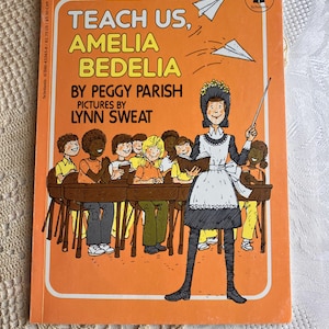 May include: A children's paperback book titled Teach Us, Amelia Bedelia by Peggy Parish with illustrations by Lynn Sweat. The orange cover features a teacher standing in front of a classroom of students with two paper airplanes flying above.