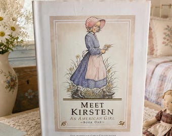 May include: A hardcover book titled Meet Kirsten: An American Girl, Book One from The American Girls Collection. The cover features a color illustration of a young person in a blue dress, pink apron, and bonnet, standing in a field of flowers.