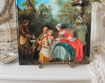 May include: A hardcover book titled The History of the Family A Record Book, featuring a colorful oil painting on the front cover depicting a group of people in 18th-century attire gathered in a garden setting with trees and a stone urn.