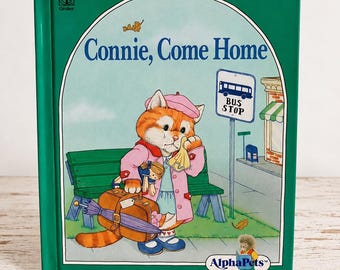 May include: A green hardcover children's book titled Connie, Come Home from the AlphaPets series. The cover illustration features an orange cat wearing a pink coat and beret, holding a brown suitcase and a doll while standing at a bus stop.