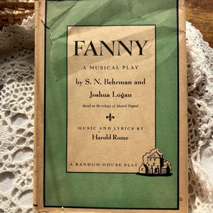 May include: A vintage copy of the musical play "FANNY" by S. N. Behrman and Joshua Logan. The book cover features a green border and a central panel with the title in bold black letters. The cover also includes the author's names and the phrase "A Random House Play."