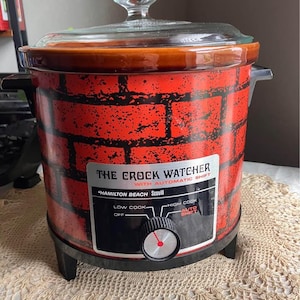 May include: A Hamilton Beach Scovill The Crock Watcher slow cooker with a red brick pattern exterior. It features a black control panel with a dial for Off, Low Cook, High Cook, and Auto Shift settings, topped with a clear glass lid and handle.
