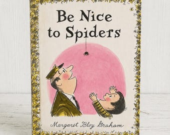 May include: A hardcover children's book titled Be Nice to Spiders by Margaret Bloy Graham. The cover features a pink circular background with a small spider hanging from a thread, observed by a man in a uniform and a child. A yellow leaf border frames the illustration.