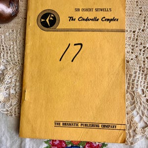 May include: A vintage, yellow-toned booklet titled "The Cinderella Complex" by Sir Osbert Sitwell. The number "17" is handwritten on the cover. The Dramatic Publishing Company is printed at the bottom. The booklet is on a lace tablecloth.