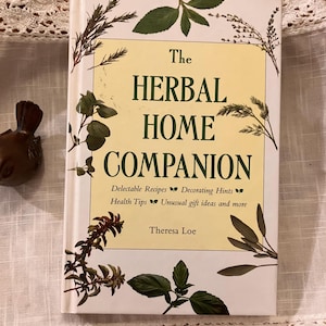 May include: A hardcover book titled "The Herbal Home Companion" with a cream-colored cover and botanical illustrations. The book features text about recipes, decorating, health tips, and gift ideas. The author's name, Theresa Loe, is also visible.