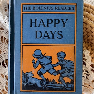 May include: A blue book with the title "The Bolenius Readers Happy Days" in black and orange lettering. The cover features an illustration of two children running with a dog in the background.