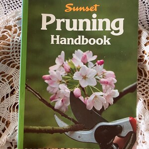 May include: A green book with the title "Sunset Pruning Handbook" in white text. The cover features a close-up of a branch of pink blossoms being pruned with gardening shears.