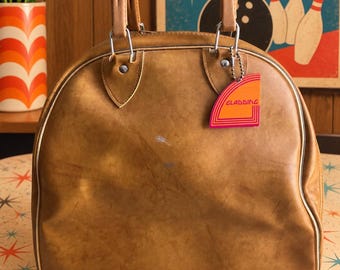 May include: A tan bowling ball bag with a rounded top, two sturdy handles, and a silver metal zipper. A small, colorful tag with the text GLADDING is attached to the handle hardware. The bag features a smooth, worn surface with light scuff marks.